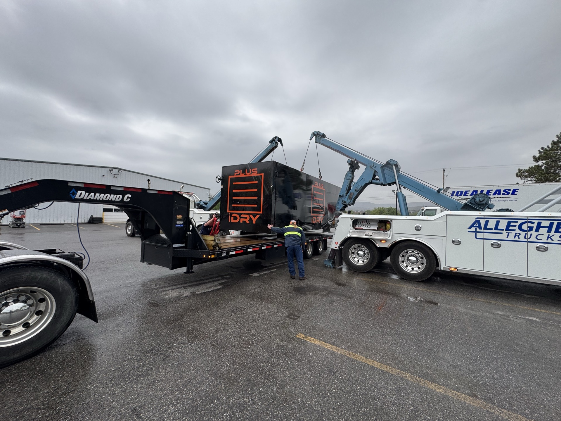 Two boom trucks lifting a black equipment unit onto a gooseneck flatbed trailer