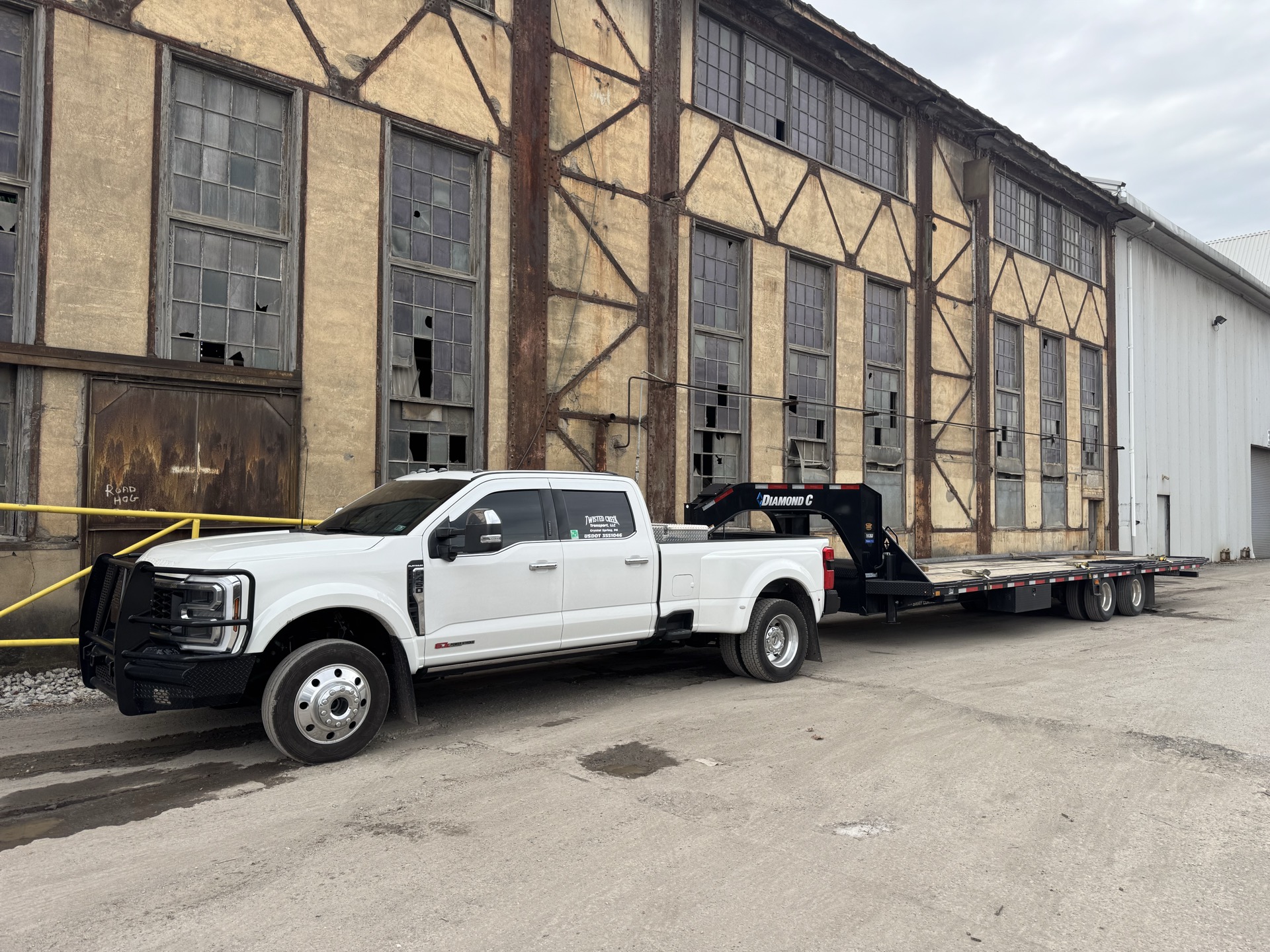 White Ford pickup with a gooseneck flatbed trailer alongside an old industrial building