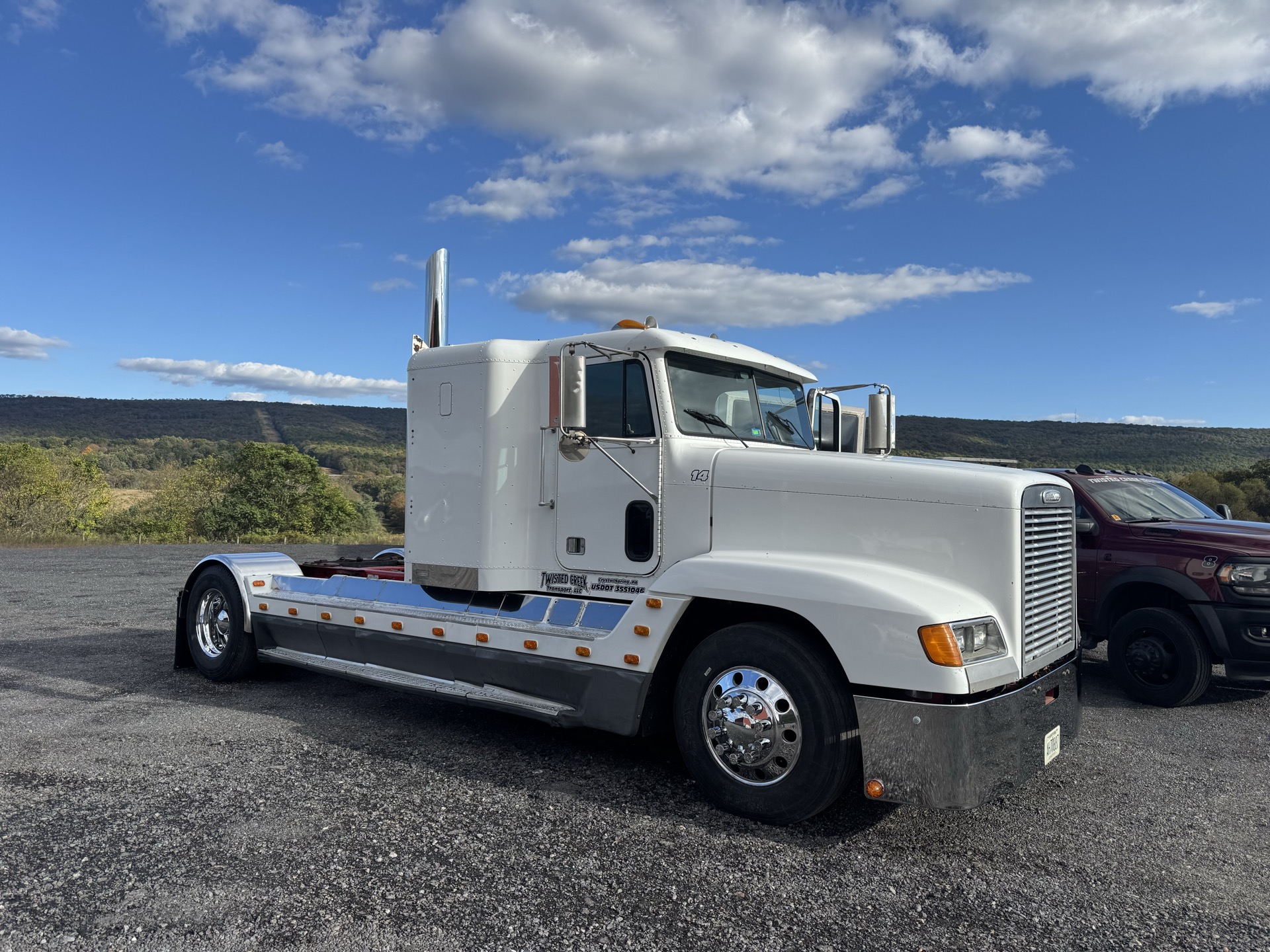 White Freightliner tractor parked on gravel with a wooded ridge behind it